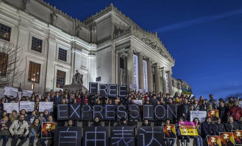 A group of people gathers in front of an illuminated classical building at night, holding signs with slogans. Prominent signs read Free Expression in English and Chinese. Other signs include various messages, held by individuals in the crowd.