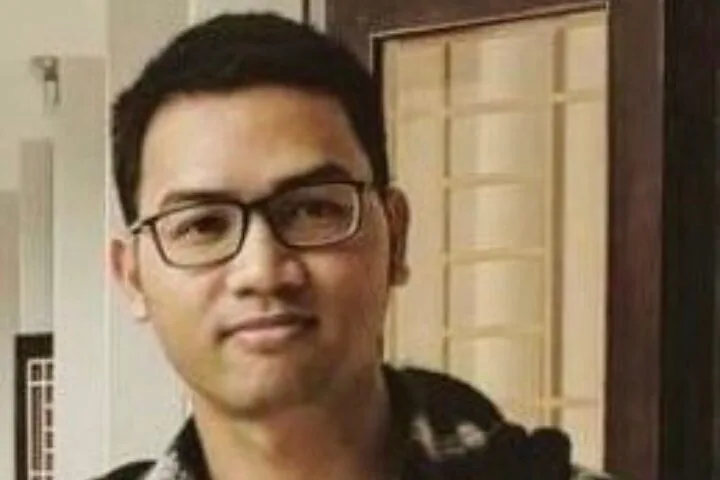 A person with short dark hair and glasses is standing indoors, wearing a dark jacket over a checkered shirt. The background includes a door with glass panels.